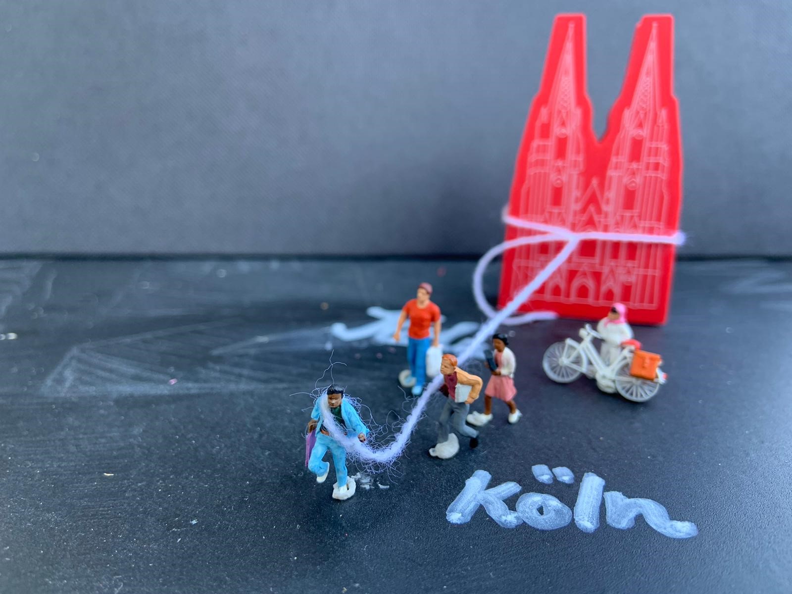 Figuren sind durch einen Faden mit einem Symbolbild des Kölner Doms verbunden.