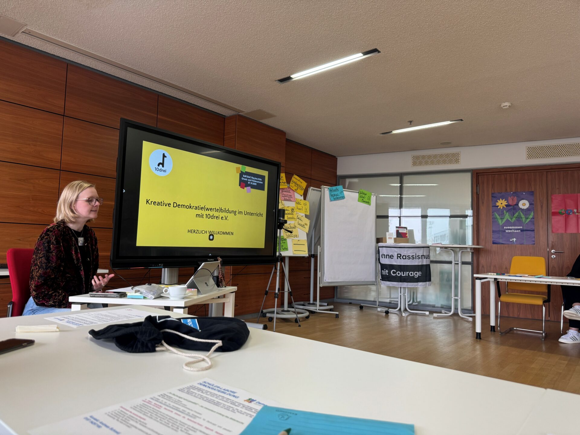 Eine Frau sitzt an einem Tisch und hält vor einer Gruppe einen Vortrag. Auf einem großen Bildschirm ist eine deutschsprachige Präsentation mit dem Titel Kreative Demokratiebildung im Unterricht zu sehen.