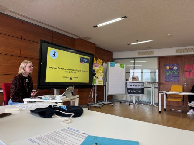 Eine Frau sitzt an einem Tisch und hält vor einer Gruppe einen Vortrag. Auf einem großen Bildschirm ist eine deutschsprachige Präsentation mit dem Titel Kreative Demokratiebildung im Unterricht zu sehen.