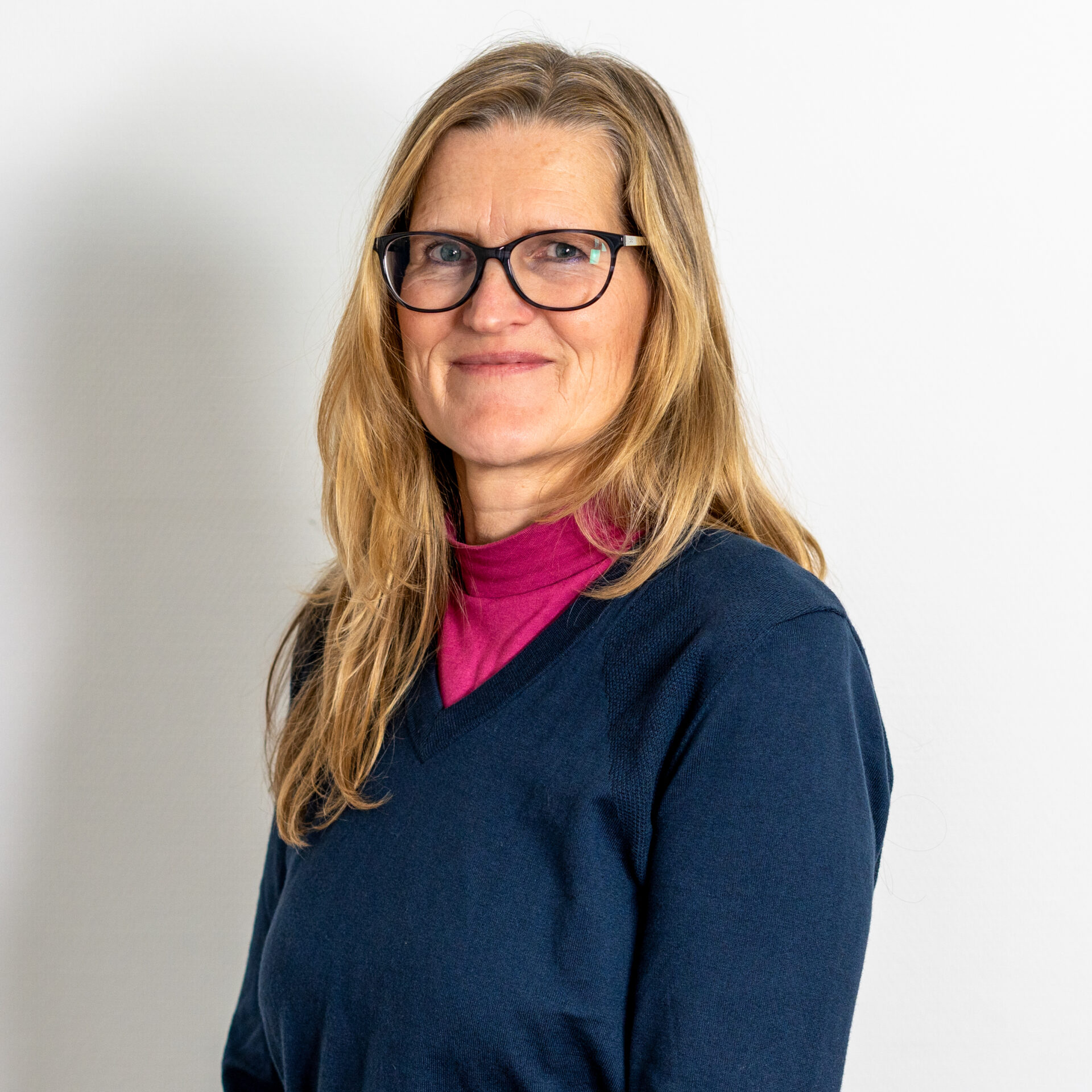 Eine Frau mit langen blonden Haaren und Brille lächelt. Sie trägt einen dunkelblauen Pullover über einem rosa Rollkragenpullover. Der Hintergrund ist schlicht weiß.