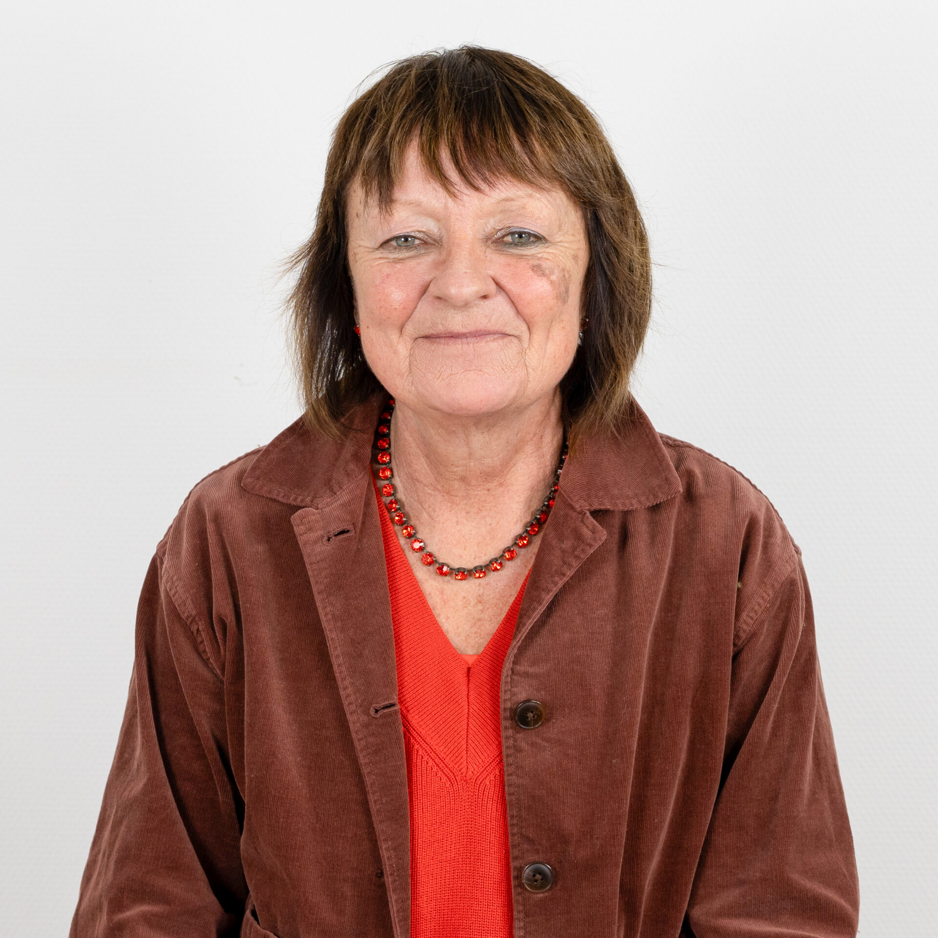 Eine Frau mit kurzen braunen Haaren, einem roten Oberteil und einer braunen Cordjacke. Sie trägt eine Perlenkette und lächelt leicht vor einem schlichten weißen Hintergrund.