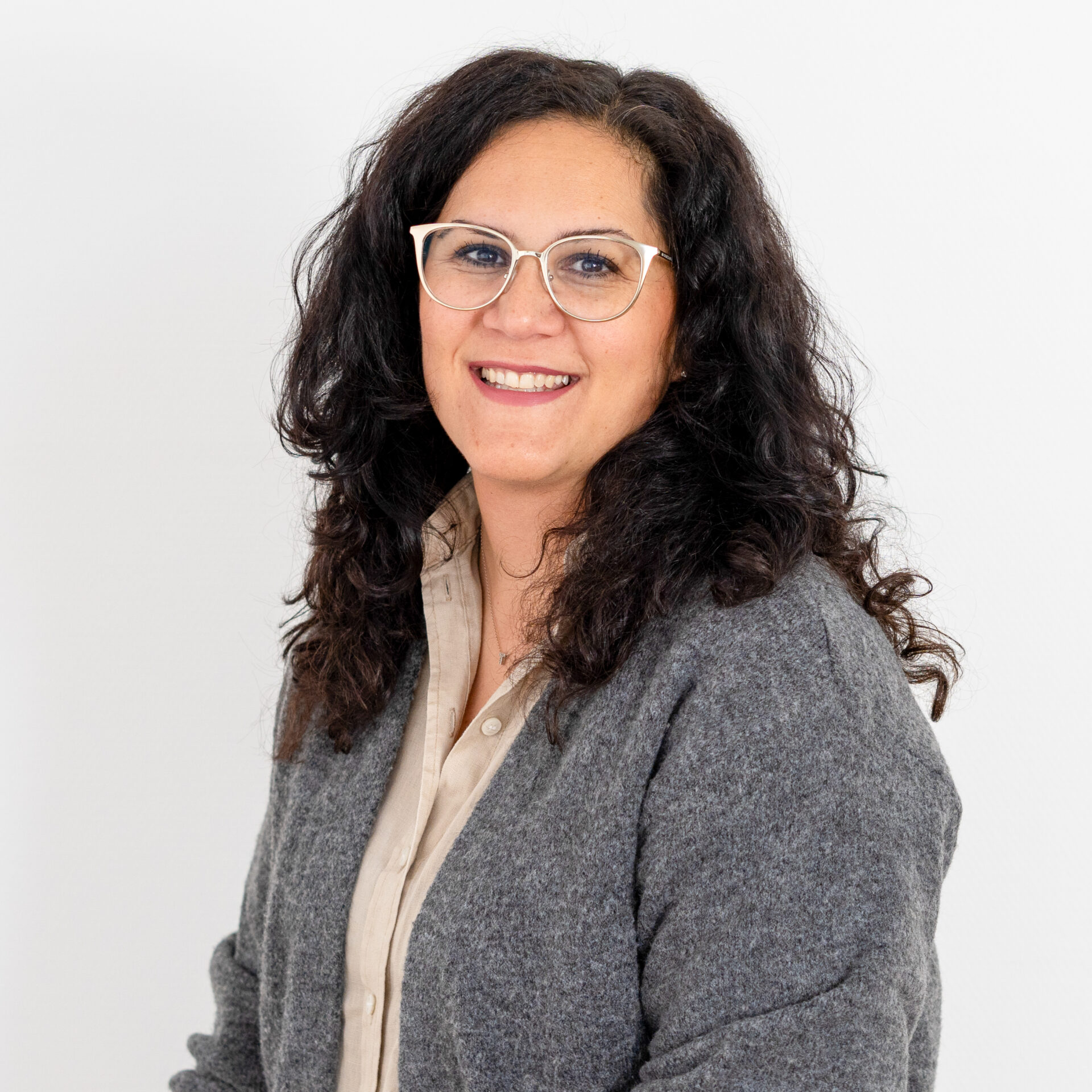 Eine Person mit lockigem schwarzem Haar und Brille, die einen grauen Pullover über einem beigen Hemd trägt, lächelt vor einem schlichten weißen Hintergrund in die Kamera.