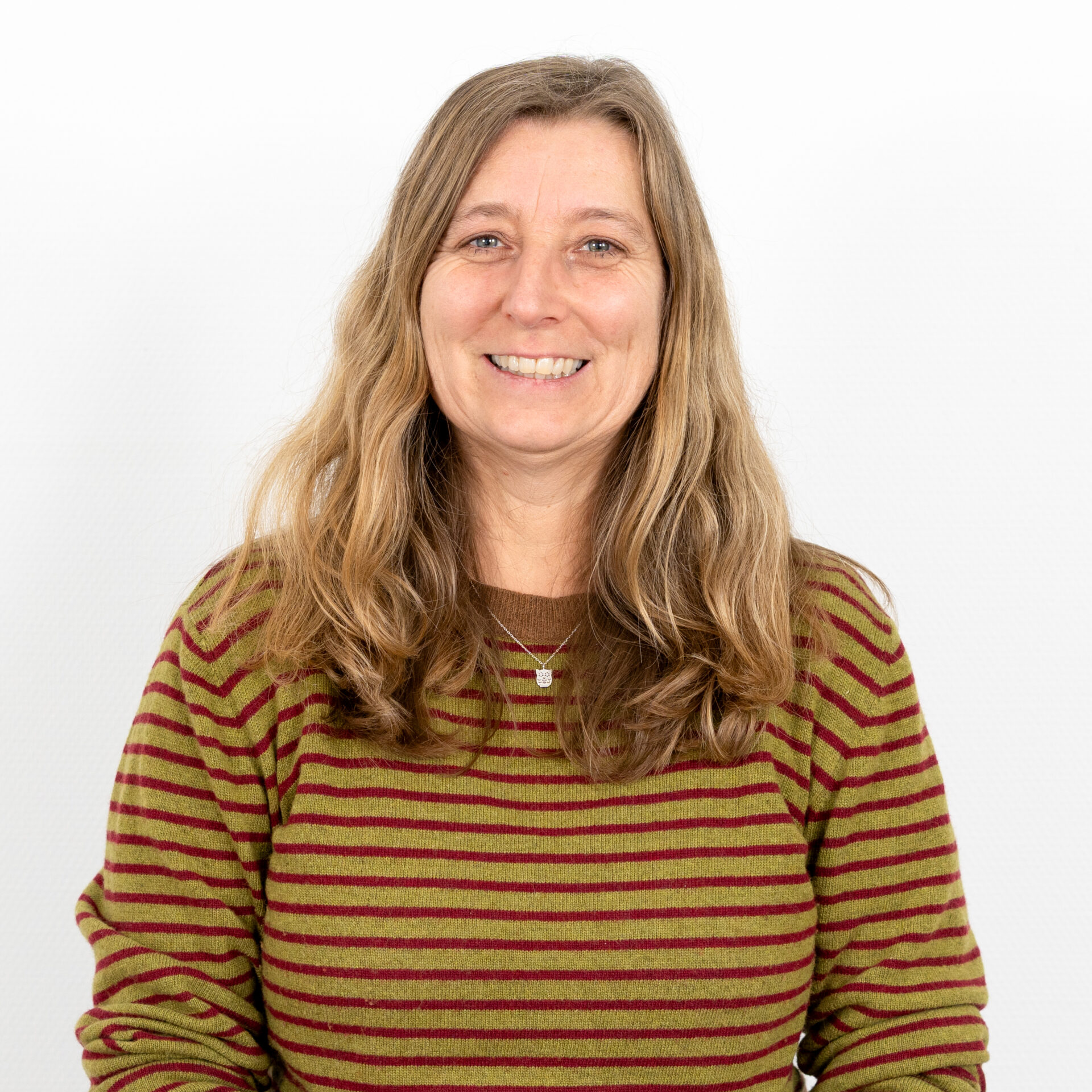Eine Person mit langen, welligen Haaren lächelt in die Kamera. Sie trägt einen grün-burgunderfarbenen gestreiften Pullover und eine Halskette. Der Hintergrund ist schlicht weiß.