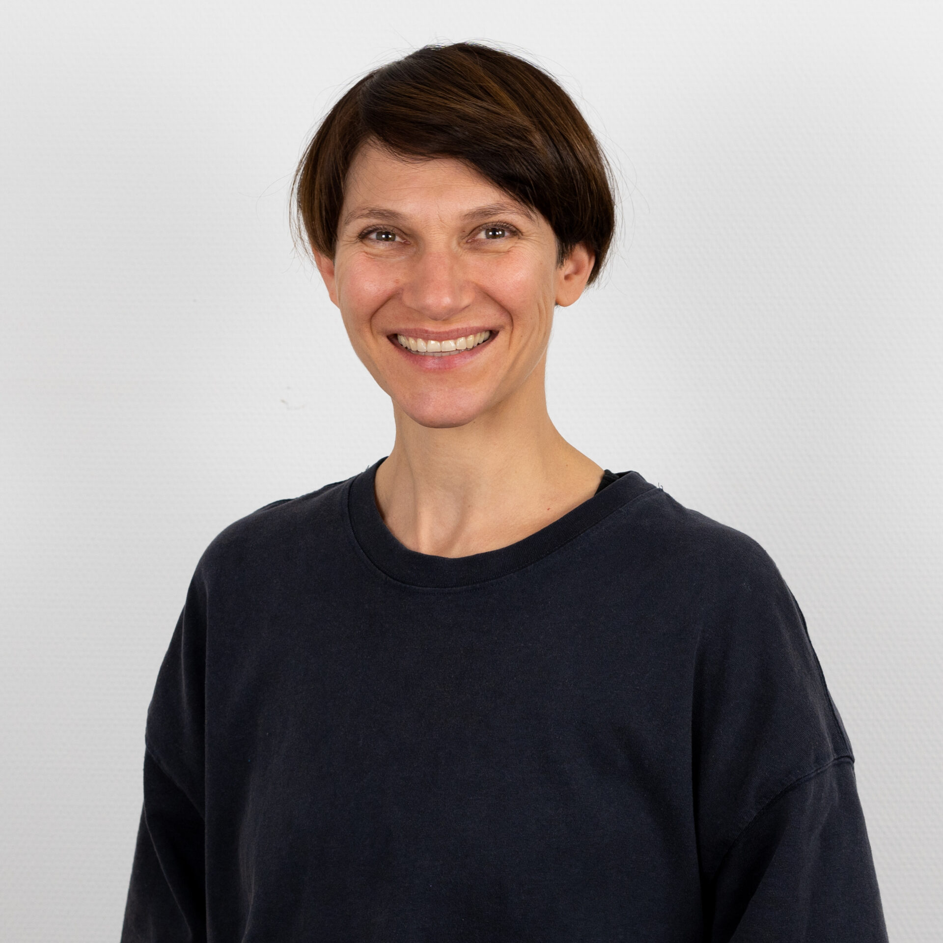 Eine Person mit kurzen braunen Haaren lächelt in die Kamera. Sie trägt ein dunkles Langarmhemd vor einem schlichten weißen Hintergrund.
