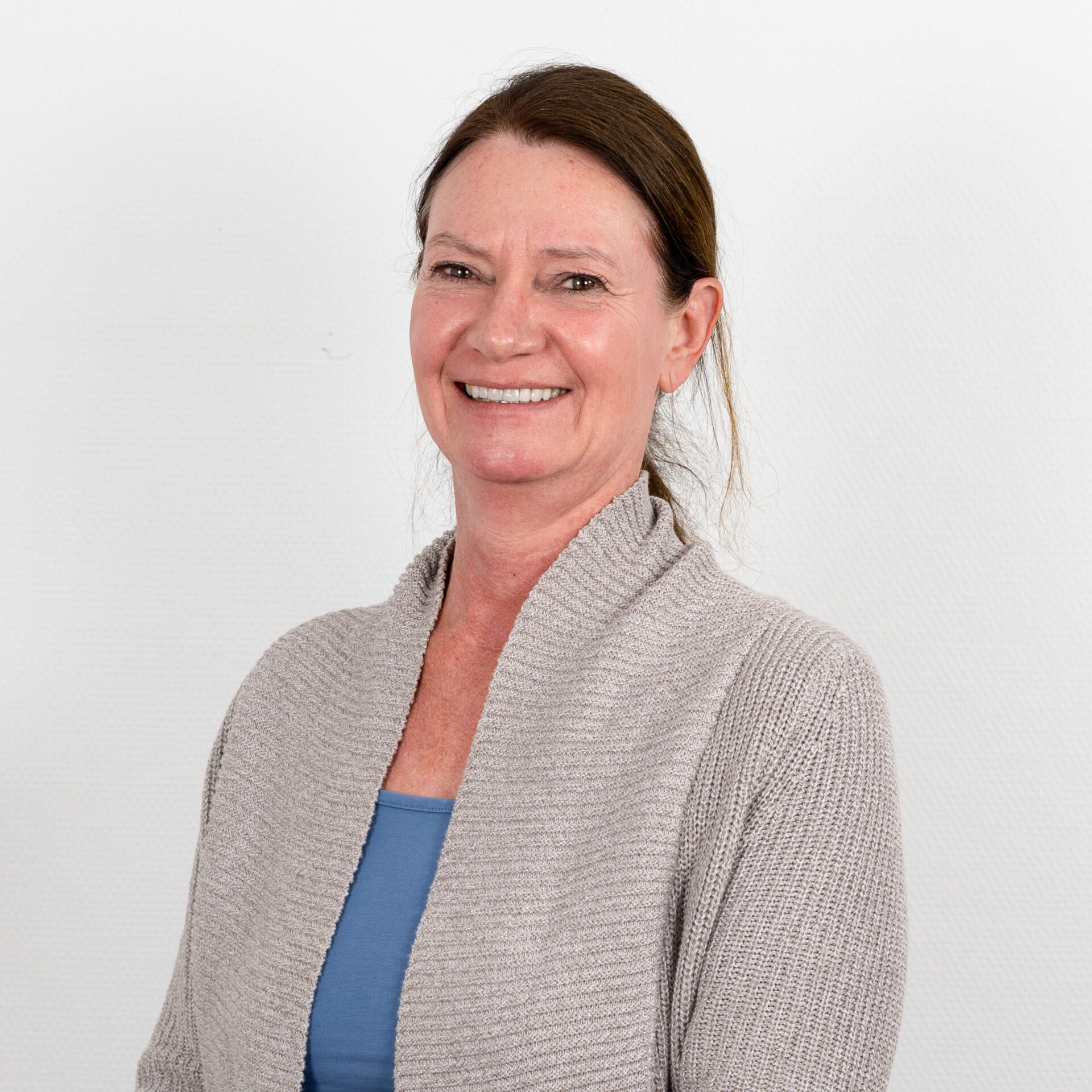 Lächelnde Frau mit zurückgebundenem braunem Haar, hellgrauer Strickjacke über blauem Oberteil, vor schlichtem weißem Hintergrund.