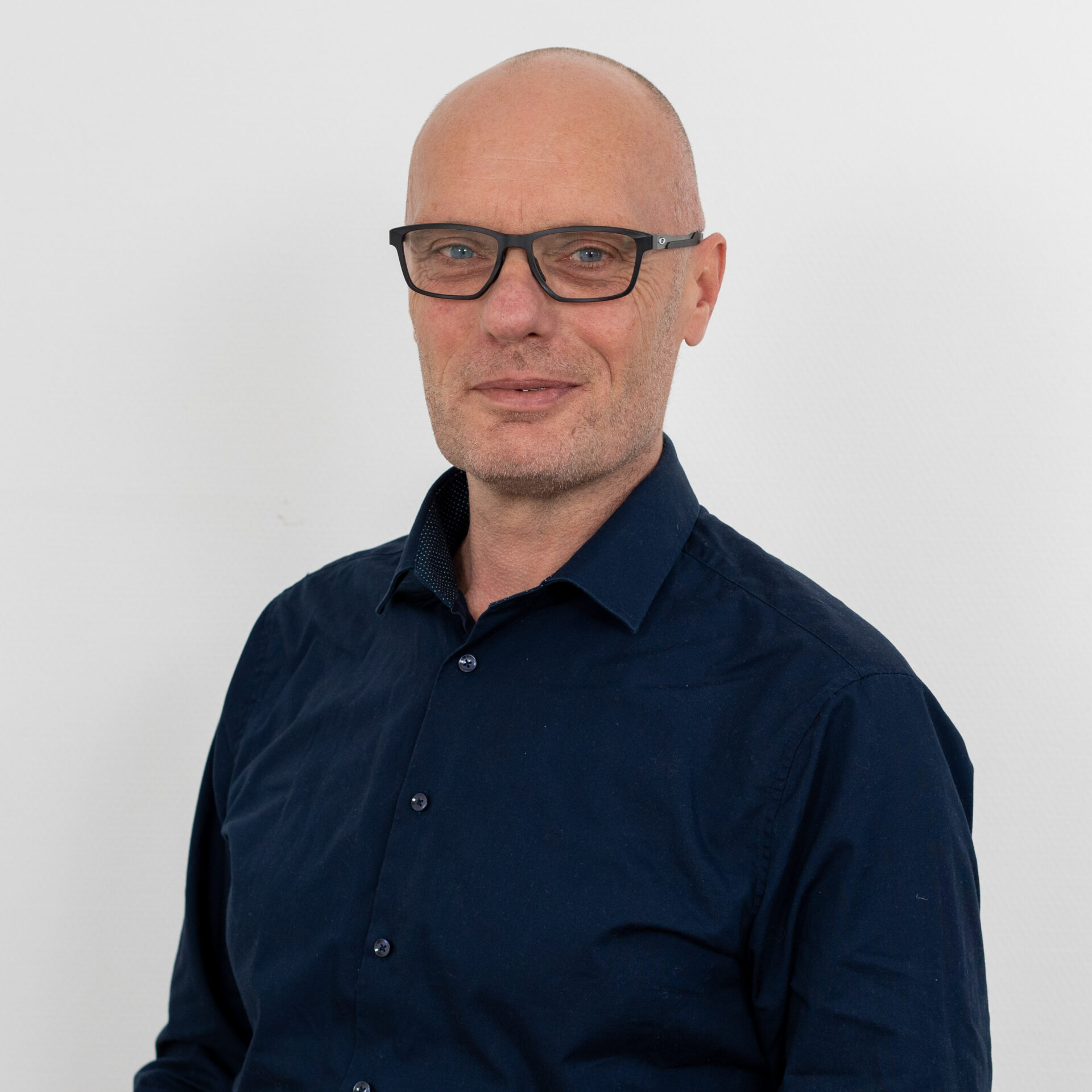 Ein Mann mit Brille und dunkelblauem Hemd steht vor einem schlichten weißen Hintergrund. Er hat einen neutralen Gesichtsausdruck und eine Glatze.