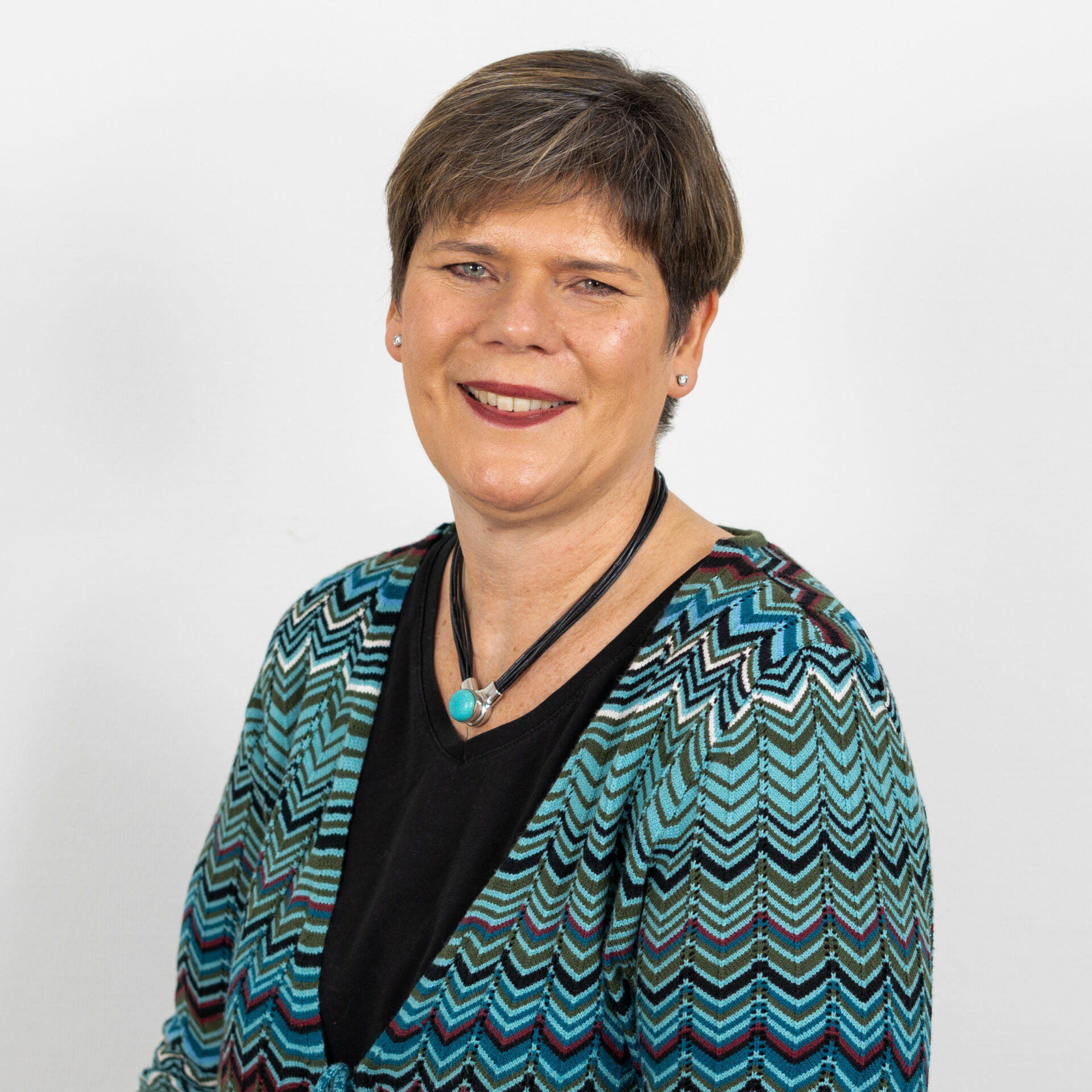 Eine Person mit kurzen Haaren lächelt und trägt eine Strickjacke mit Chevron-Muster in Blau-, Grün- und Schwarztönen über einem schwarzen Oberteil. Sie trägt eine Halskette mit einem türkisfarbenen Anhänger. Der Hintergrund ist schlicht weiß.