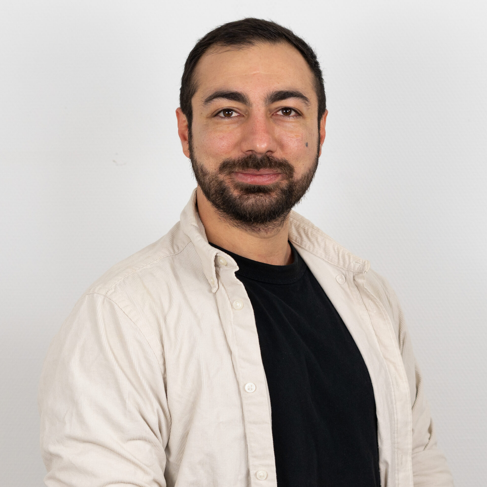 Ein bärtiger Mann mit kurzen dunklen Haaren in einem schwarzen Hemd und einem hellen Button-Up posiert vor einem weißen Hintergrund und blickt mit einem subtilen Lächeln in die Kamera.