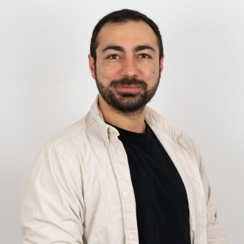 Ein bärtiger Mann mit kurzen dunklen Haaren in einem schwarzen Hemd und einem hellen Button-Up posiert vor einem weißen Hintergrund und blickt mit einem subtilen Lächeln in die Kamera.