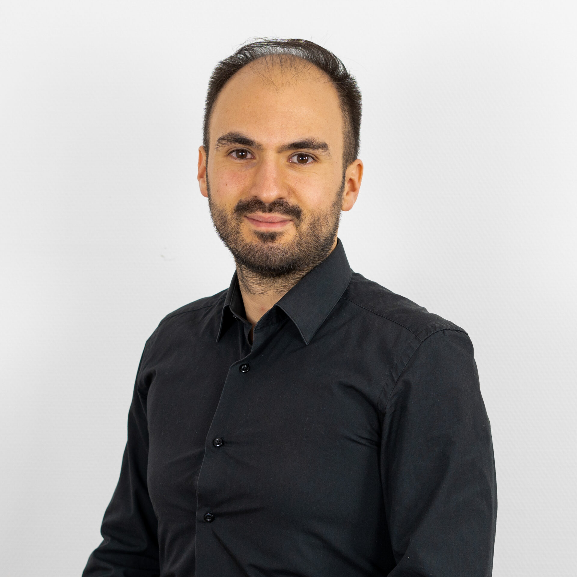 Ein Mann mit kurzen Haaren und Bart steht vor einem schlichten weißen Hintergrund. Er trägt ein schwarzes Hemd mit Knöpfen. Er lächelt leicht und blickt direkt in die Kamera.