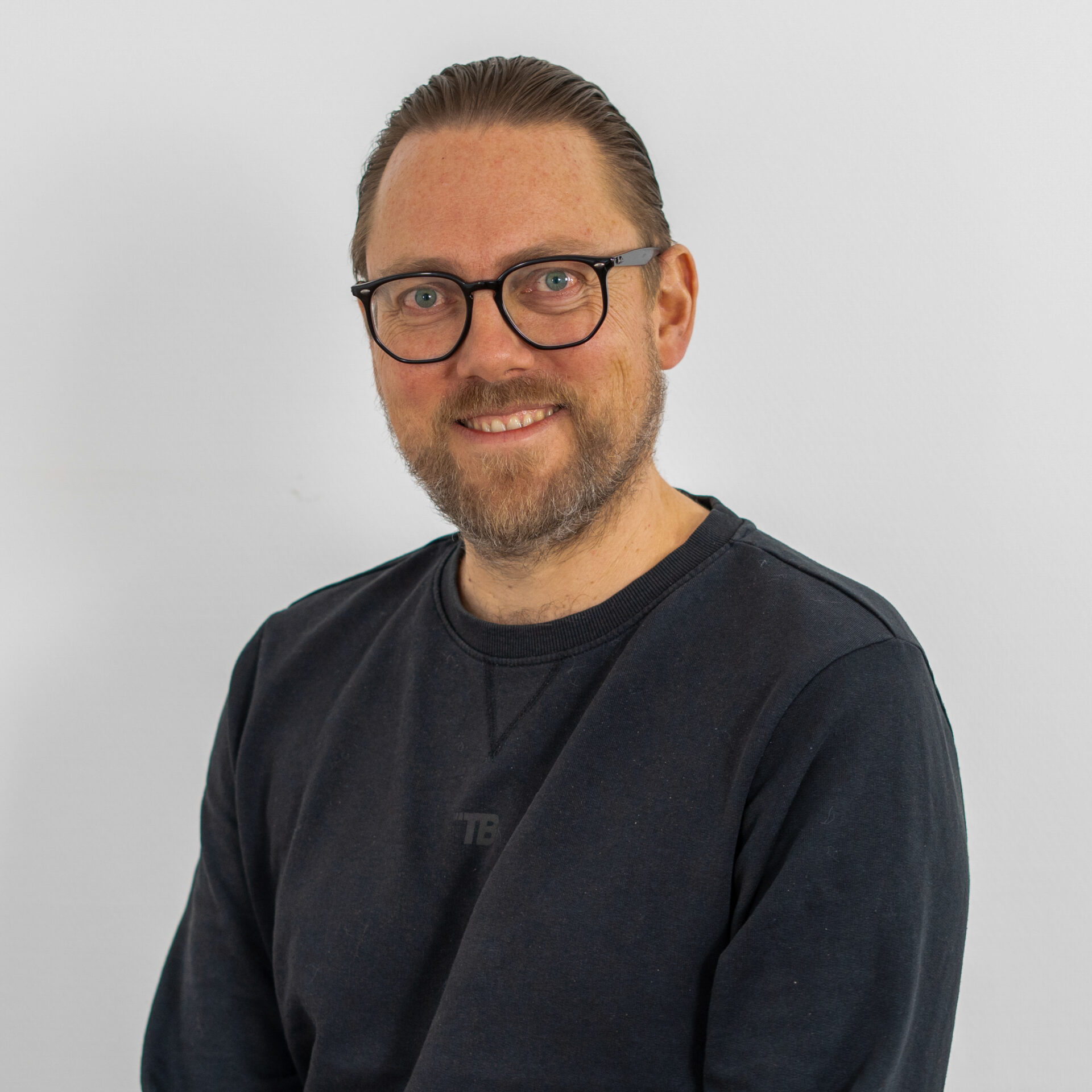 Ein Mann mit Brille und Bart lächelt in die Kamera. Er trägt einen schwarzen Pullover und hat seine Haare zurückgekämmt. Der Hintergrund ist schlicht weiß.