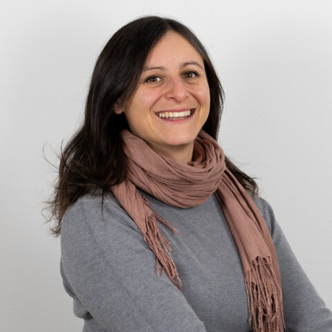 Frau mit langem dunklem Haar und einem breiten Lächeln, trägt einen grauen Pullover und einen hellrosa Schal und steht vor einem schlichten weißen Hintergrund.