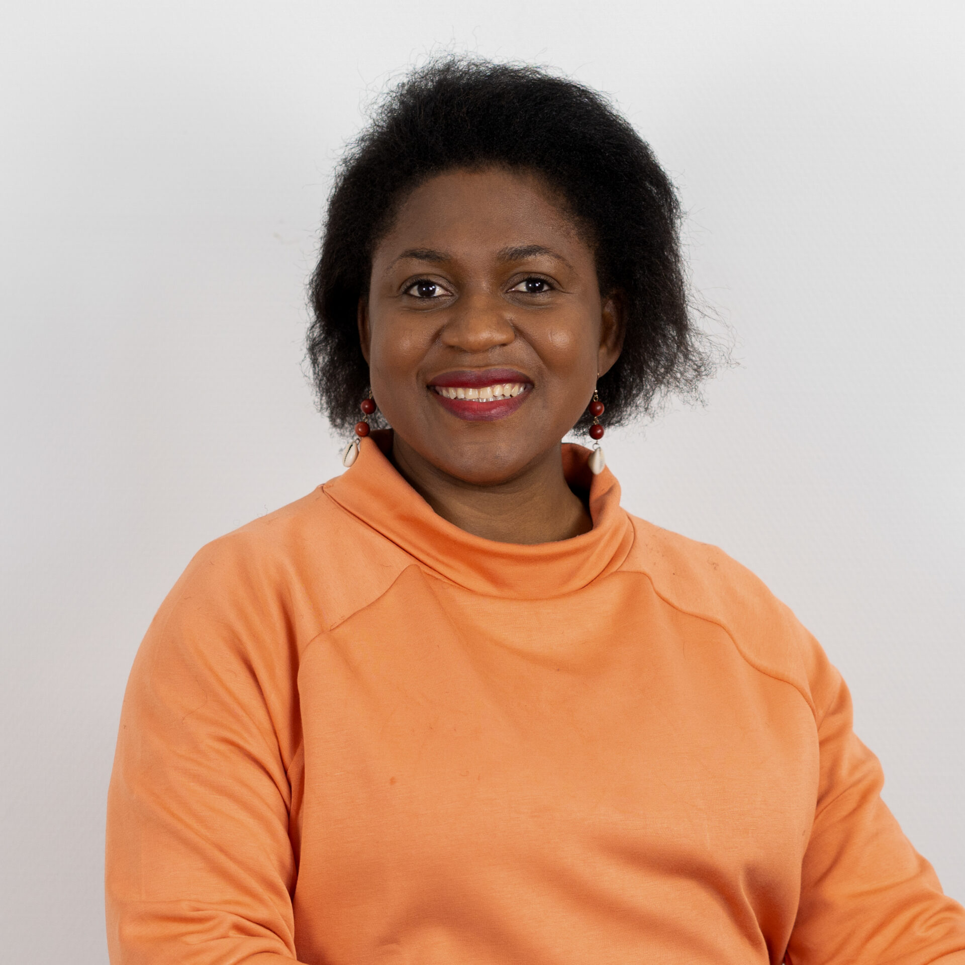 Eine Person mit kurzen lockigen Haaren lächelt in die Kamera. Sie trägt ein orangefarbenes Oberteil und rote Ohrringe. Der Hintergrund ist schlicht weiß.