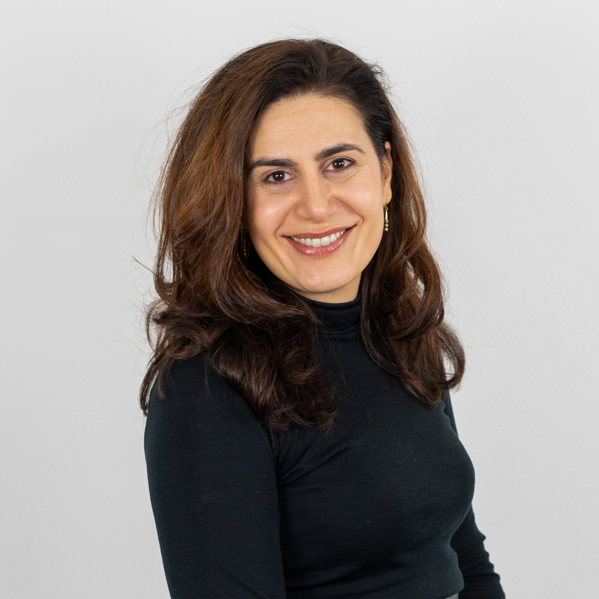 Eine Frau mit langen braunen Haaren und einem schwarzen Rollkragenpullover lächelt in die Kamera und steht vor einem schlichten weißen Hintergrund.