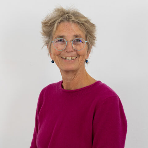 Eine ältere Person mit kurzen, zerzausten Haaren, einer Brille und einem leuchtend rosa Pullover lächelt vor einem schlichten weißen Hintergrund in die Kamera.