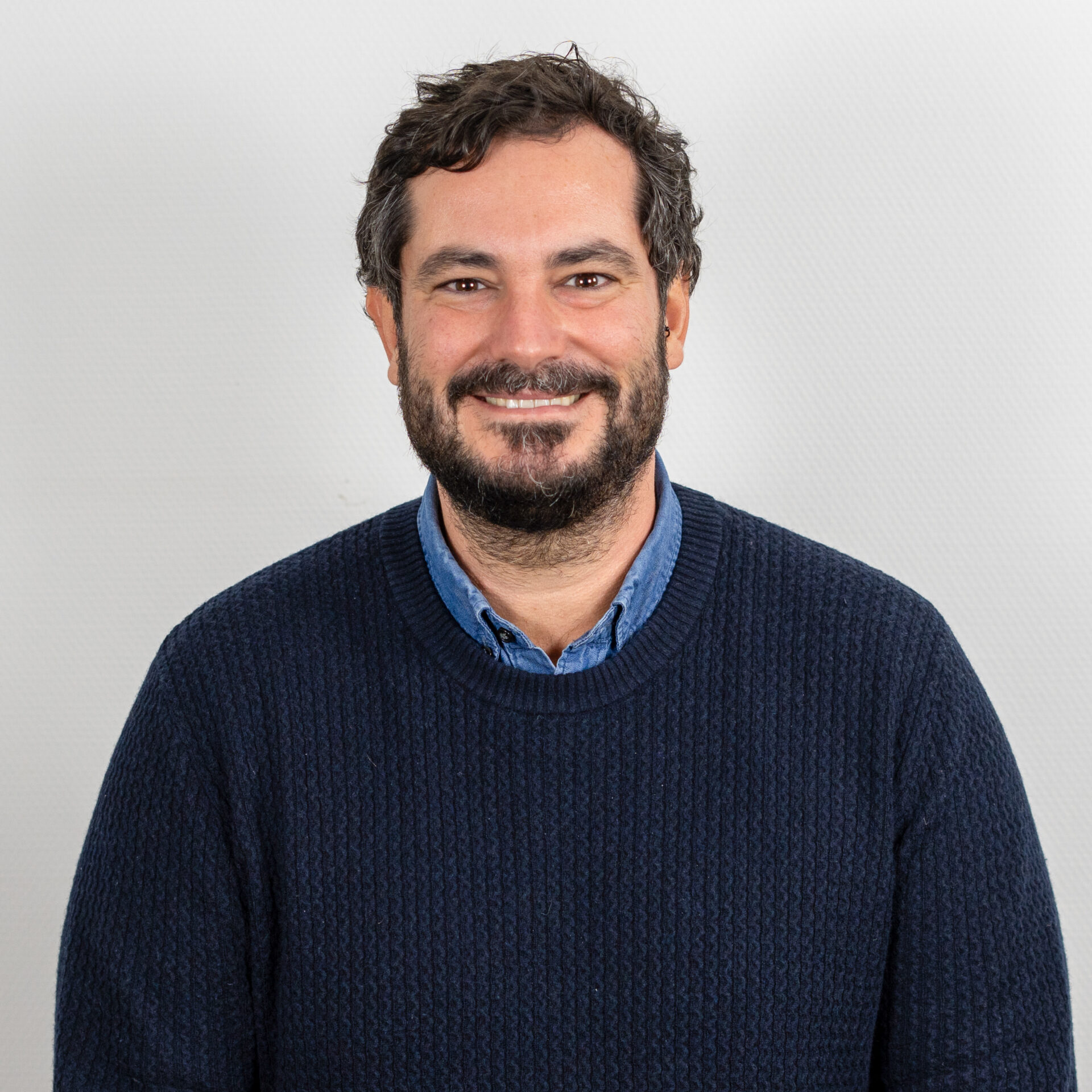 Ein Mann mit kurzen, dunklen Haaren und Bart, der einen blauen Strickpullover und ein blaues Hemd mit Kragen trägt, lächelt vor einem hellgrauen Hintergrund in die Kamera.