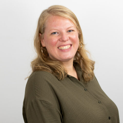 Eine Person mit schulterlangem blondem Haar lächelt in die Kamera. Sie trägt ein dunkelgrünes Hemd mit Knöpfen und Creolen und steht vor einem schlichten weißen Hintergrund.