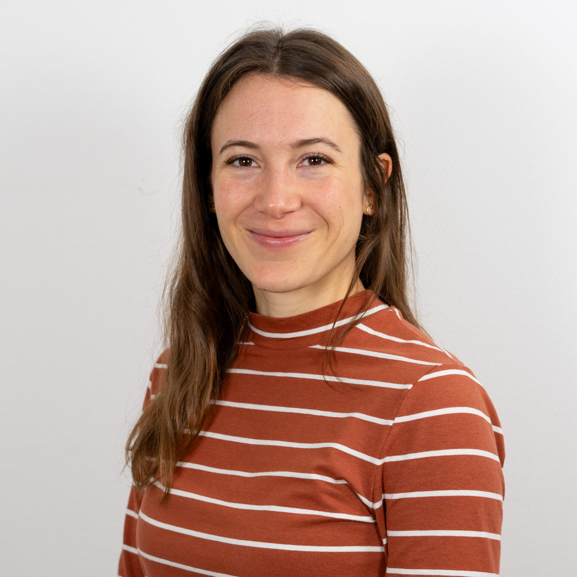Eine Person mit langen braunen Haaren lächelt in die Kamera. Sie trägt ein rostfarbenes, gestreiftes Oberteil. Der Hintergrund ist schlicht weiß.