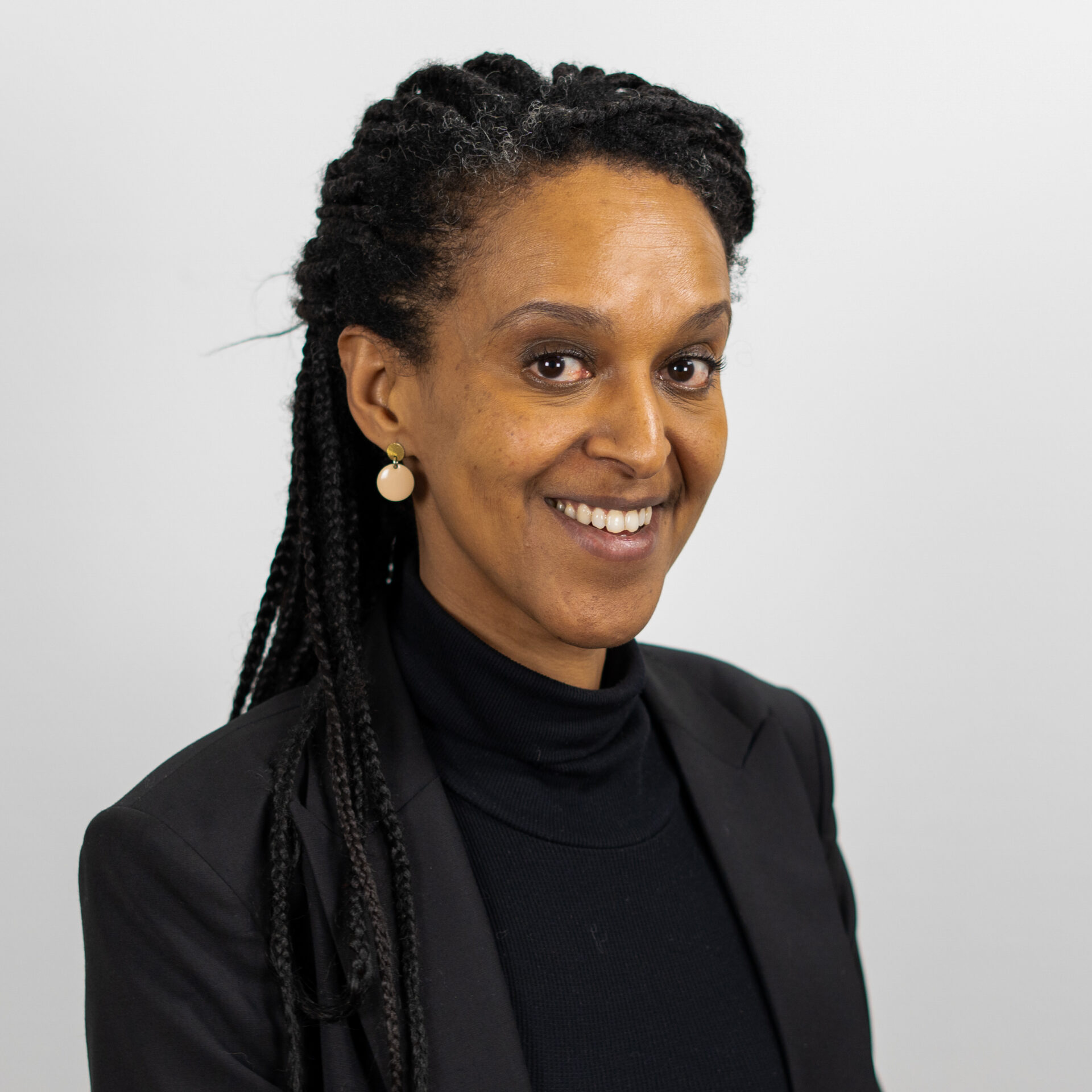 Eine Person mit geflochtenen Haaren trägt einen schwarzen Rollkragenpullover und einen schwarzen Blazer. Sie hat goldene Ohrringe und lächelt vor einem schlichten weißen Hintergrund in die Kamera.