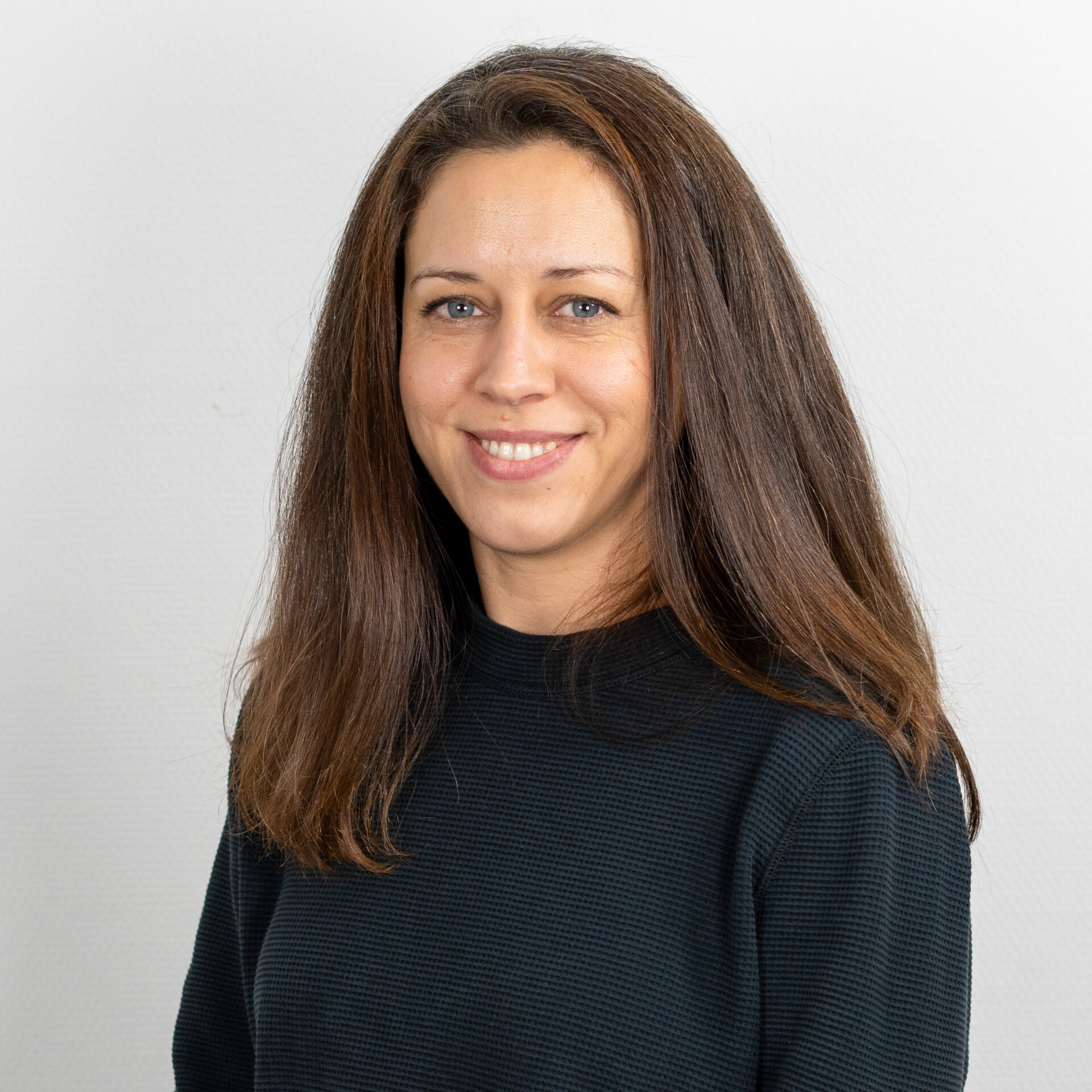 Eine lächelnde Person mit langen braunen Haaren und einem schwarzen Oberteil steht vor einem schlichten weißen Hintergrund.