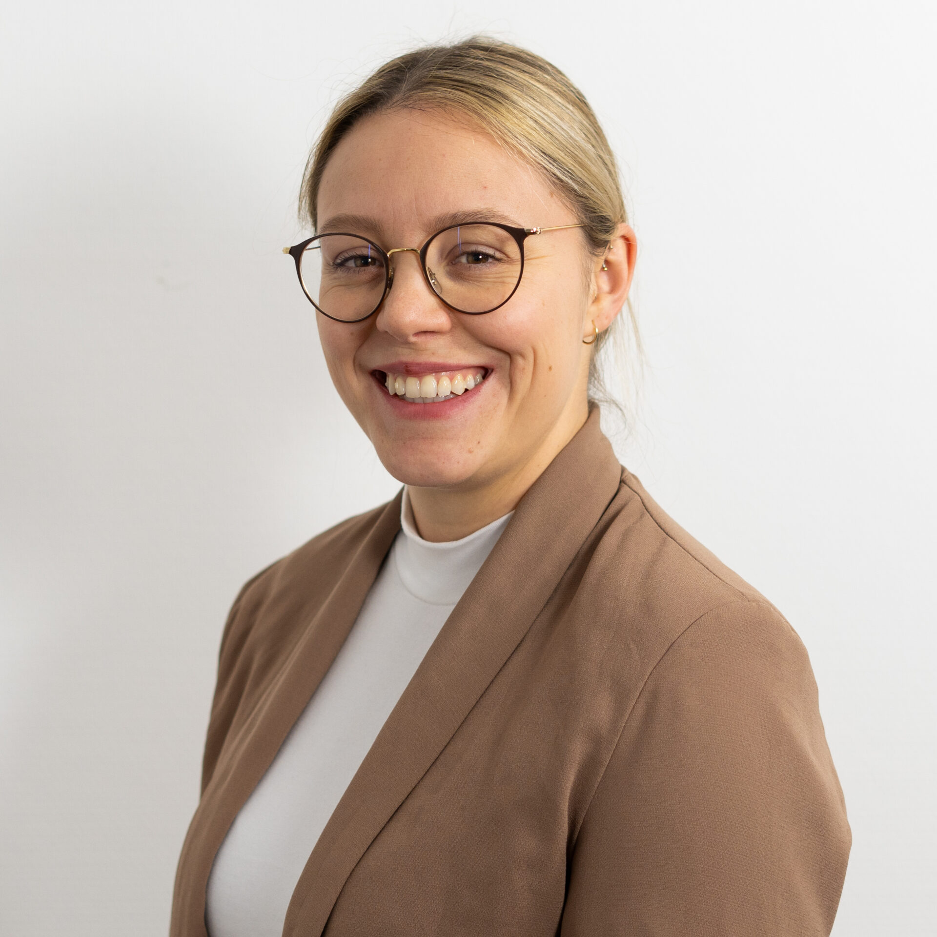 Lächelnde Person mit Brille und blondem Haar zu einem Dutt, trägt einen braunen Blazer und ein weißes Oberteil vor einem schlichten weißen Hintergrund.