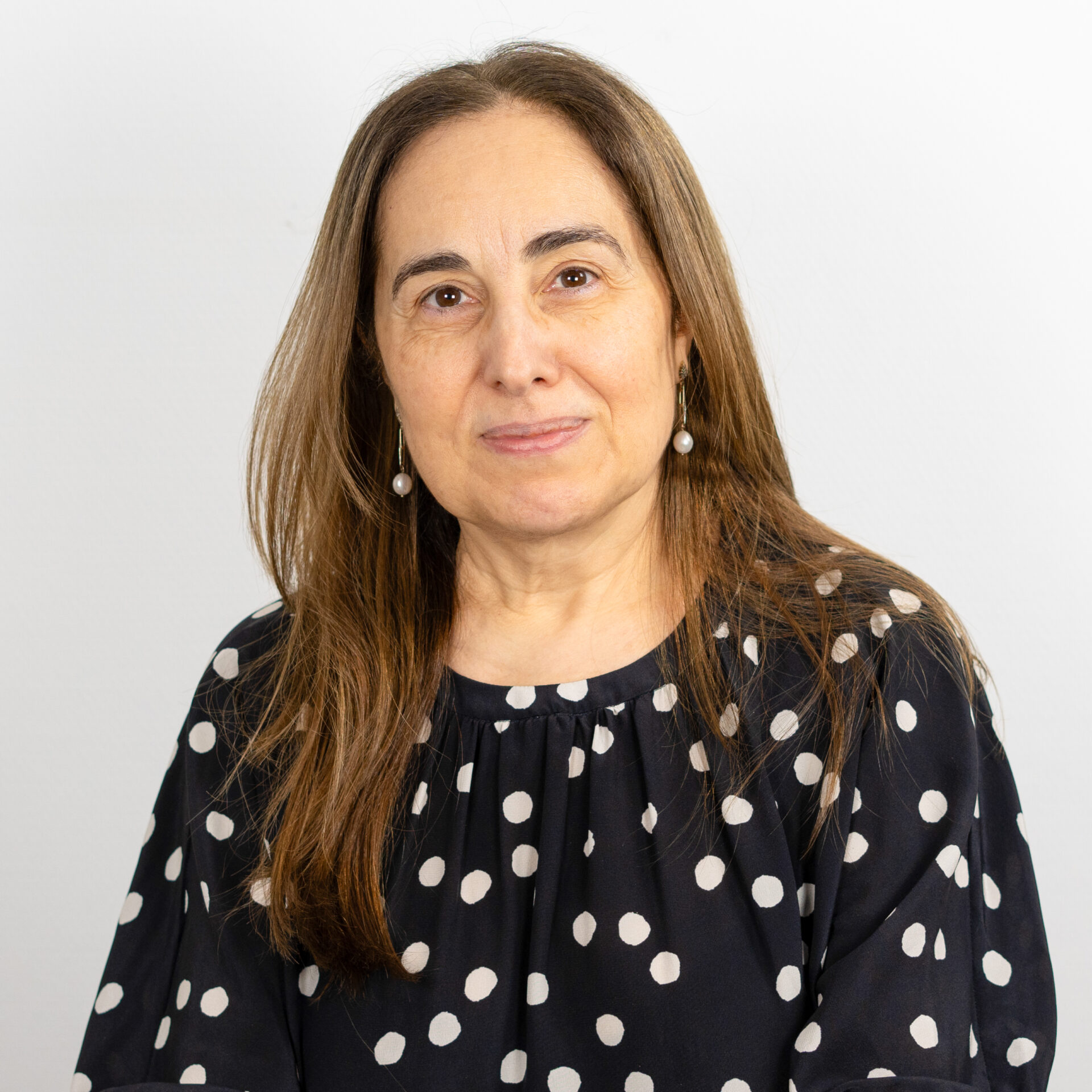 Eine Person mit langen braunen Haaren trägt eine gepunktete Bluse und Perlenohrringe und blickt mit neutralem Gesichtsausdruck in die Kamera. Der Hintergrund ist eine schlichte, helle Wand.