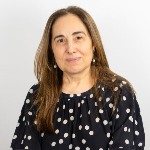 Eine Person mit langen braunen Haaren trägt eine gepunktete Bluse und Perlenohrringe und blickt mit neutralem Gesichtsausdruck in die Kamera. Der Hintergrund ist eine schlichte, helle Wand.