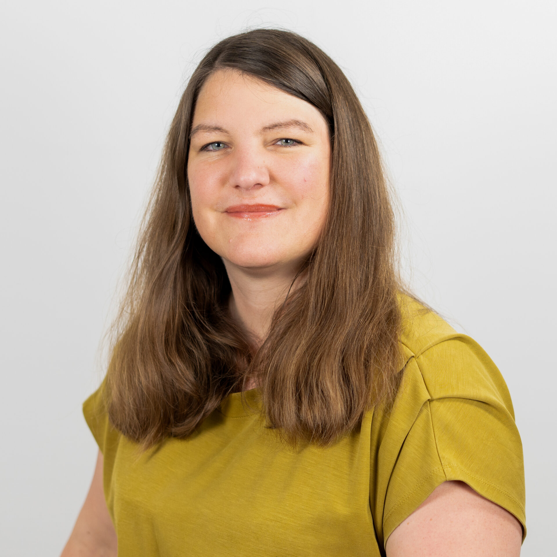 Frau mit langem braunem Haar und gelbem Hemd, die vor einem schlichten weißen Hintergrund sanft lächelt.