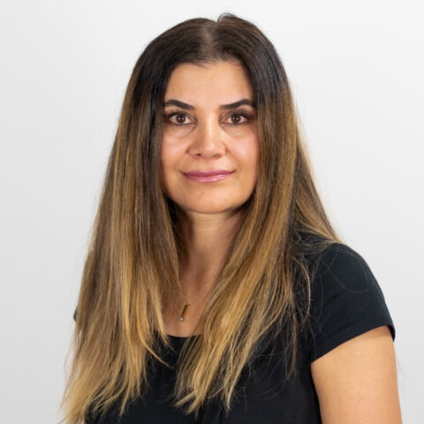 Eine Frau mit langem, glattem, dunklem Haar und einem leichten Lächeln, die ein schwarzes Oberteil trägt, posiert vor einem schlichten weißen Hintergrund.