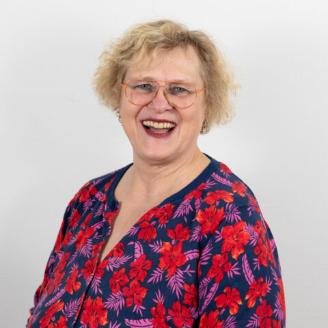 Lächelnde Frau mit kurzen lockigen Haaren, Brille und blauem Oberteil mit rotem Blumenmuster, steht vor einem schlichten weißen Hintergrund.