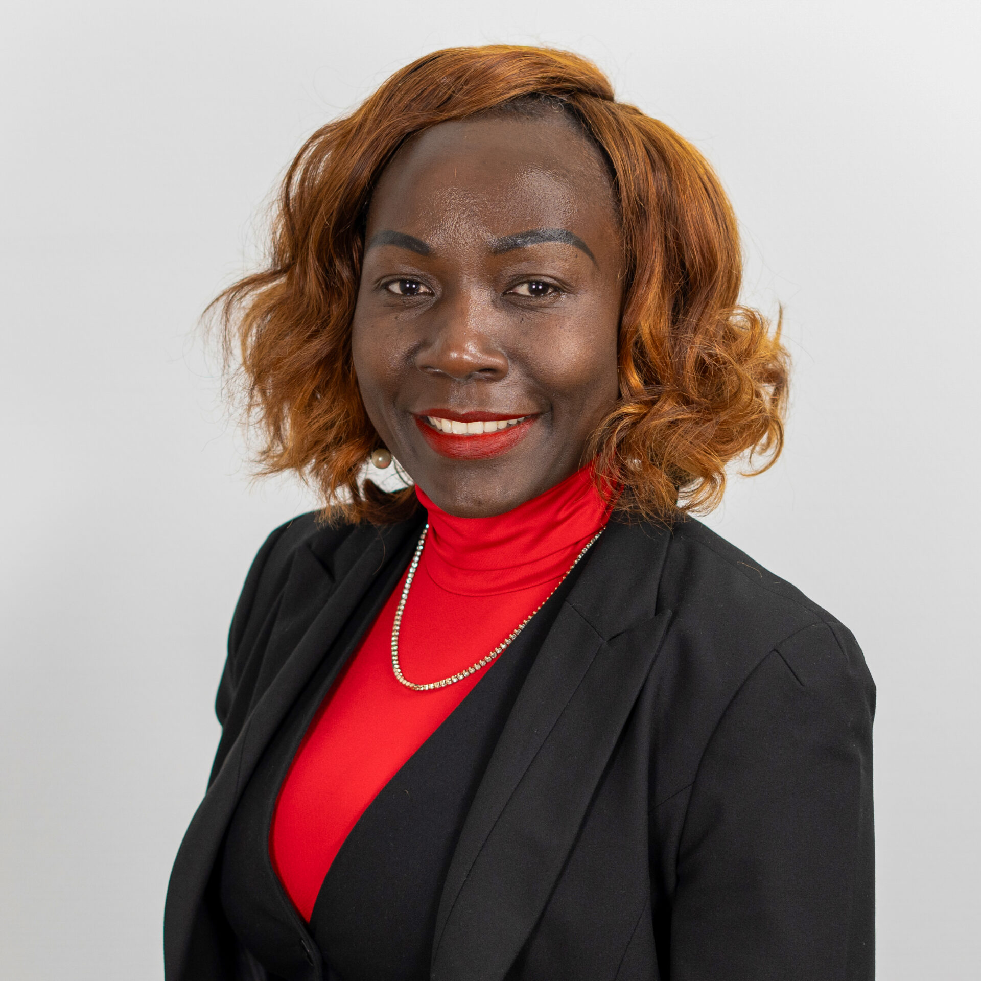 Eine Person mit schulterlangem, welligem Haar lächelt. Sie trägt einen schwarzen Blazer über einem roten Oberteil und eine silberne Halskette. Der Hintergrund ist schlicht weiß.