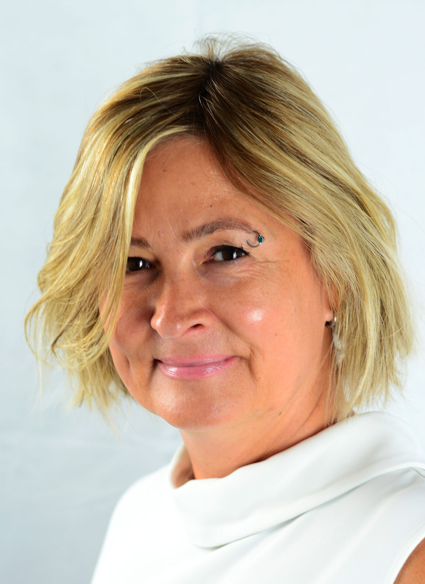 Eine Frau mit kurzen blonden Haaren und einem weißen Oberteil lächelt leicht. Sie hat ein kleines Muster neben ihrem linken Auge. Der Hintergrund ist schlicht und hell.