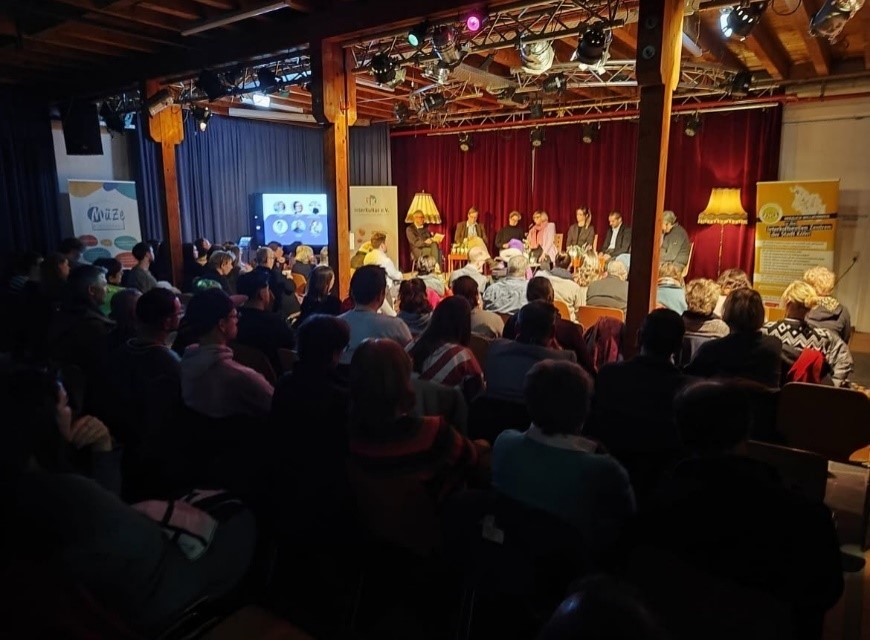 Ein großes Publikum sitzt vor einer Bühne, auf der mehrere Personen vor roten Vorhängen und warmer Beleuchtung Platz nehmen. Der Raum ist mit freiliegenden Holzbalken, Bannern und einem Monitor ausgestattet, der Bilder zeigt.