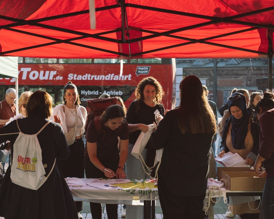 Menschen versammeln sich unter einem roten Zelt bei einer Veranstaltung im Freien, einige tragen sich ein oder holen sich Artikel von einem Tisch. Im Hintergrund ist ein roter Stadtrundfahrtbus zu sehen. Die Atmosphäre ist lebhaft und locker.
