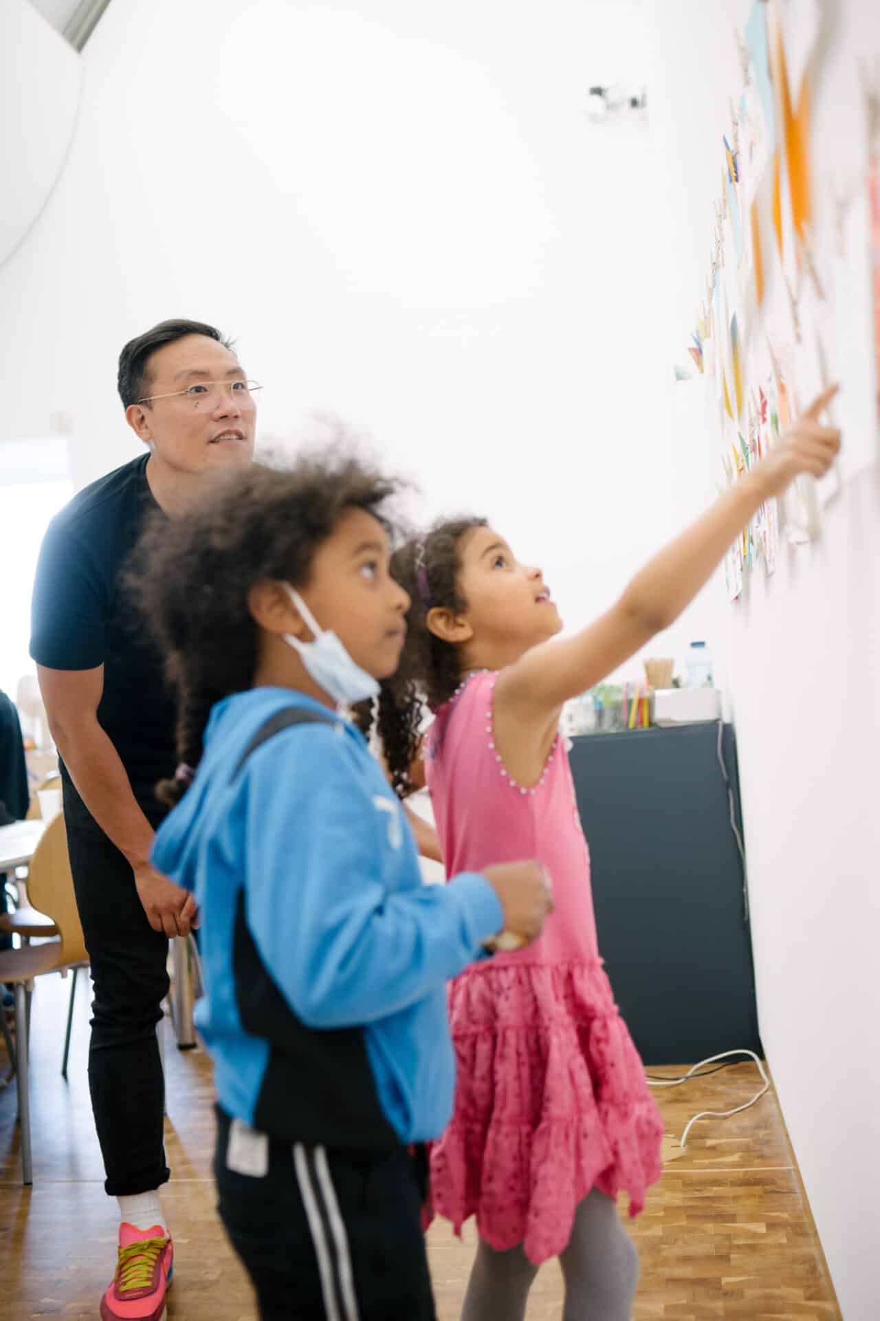 Im Vordergrund stehen zwei Kinder, eins zeigt auf ein Poster an der Wand. Ein Mann im Hintergrund schaut ebenfalls auf das Poster.