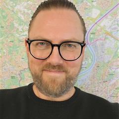 Mann mit kurzen, nach hinten gegelten Haaren, Bart und schwarzer Brille, trägt einen dunklen Pullover und lächelt vor einem bunten Stadtplan-Hintergrund.