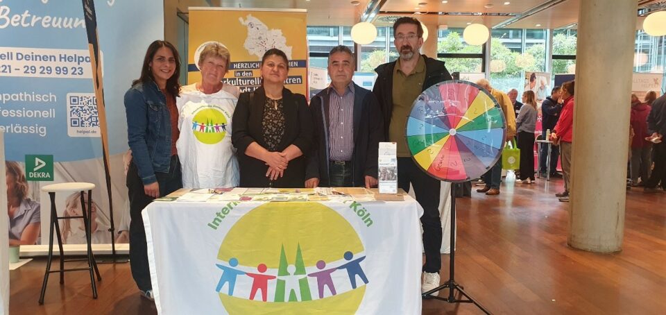 Fünf Personen stehen bei einer Indoor-Veranstaltung hinter einem Infotisch mit einem bunten Logo und Flyern; rechts ist ein Glücksrad aufgestellt. Im Hintergrund sind andere Teilnehmer und Stände zu sehen.