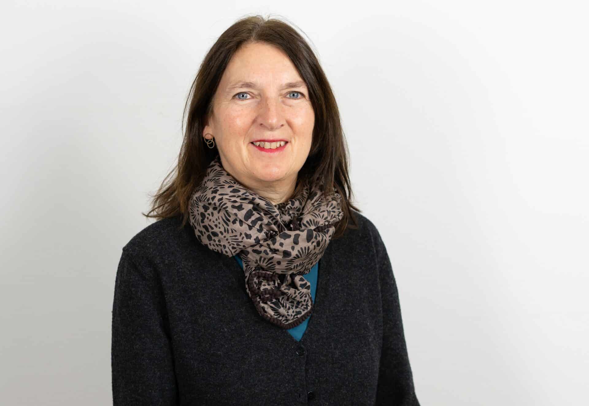 Eine Frau mit langen braunen Haaren, die eine dunkle Strickjacke und einen gemusterten Schal trägt, lächelt in die Kamera vor einem schlichten weißen Hintergrund.