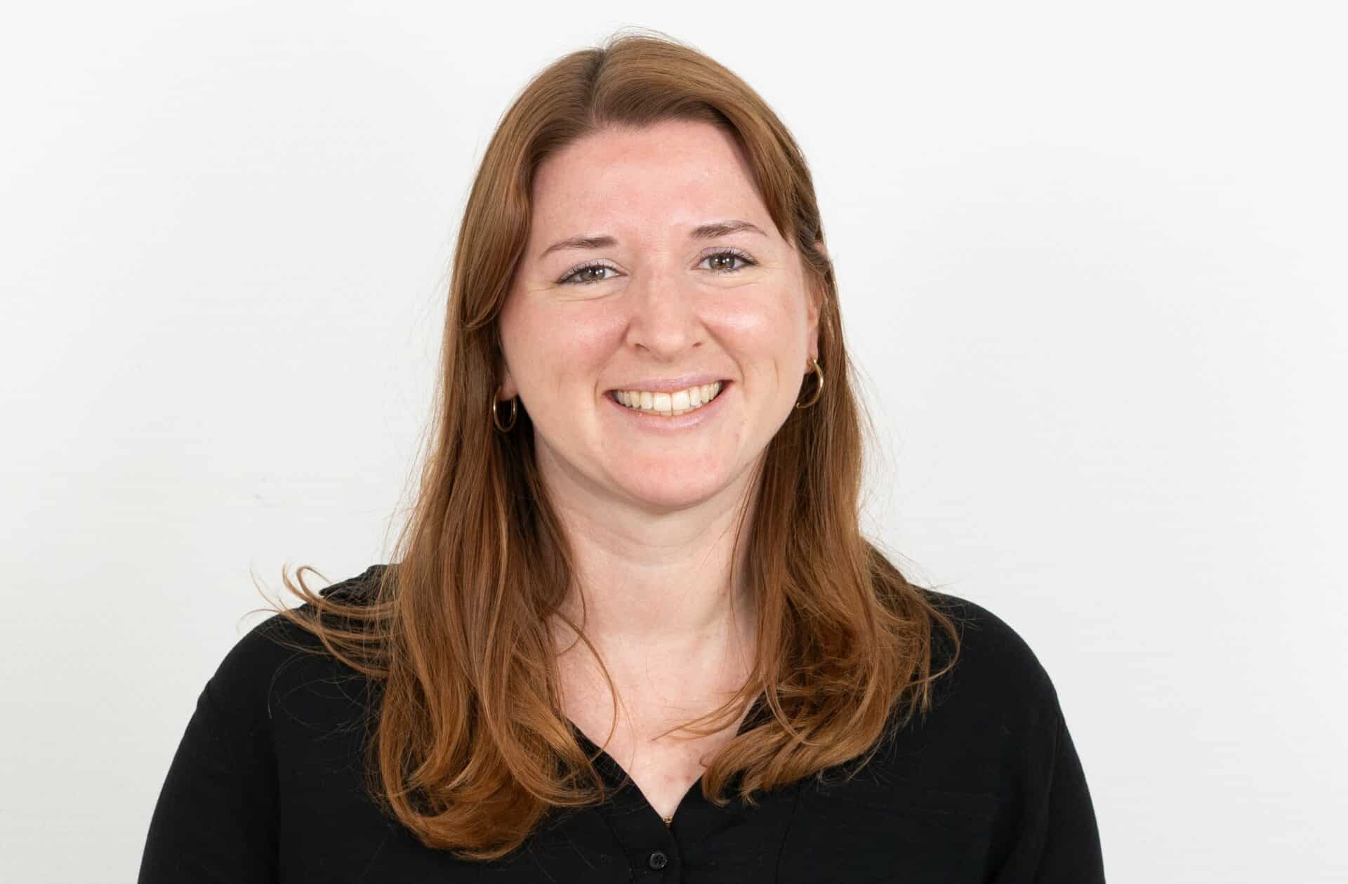 Eine Frau mit langen hellbraunen Haaren, einem schwarzen Oberteil und Ohrringen lächelt in die Kamera vor einem schlichten weißen Hintergrund.