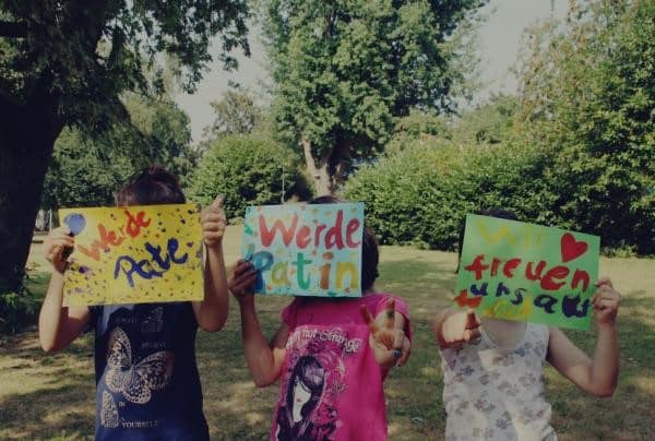 Kinder halten draußen bunte Schilder hoch, auf denen "Werde Pate" steht.
