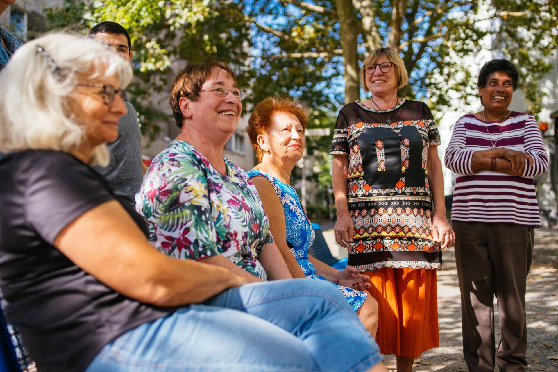 In einem Park sitzen und stehen ältere Damen in einem Halbkreis.