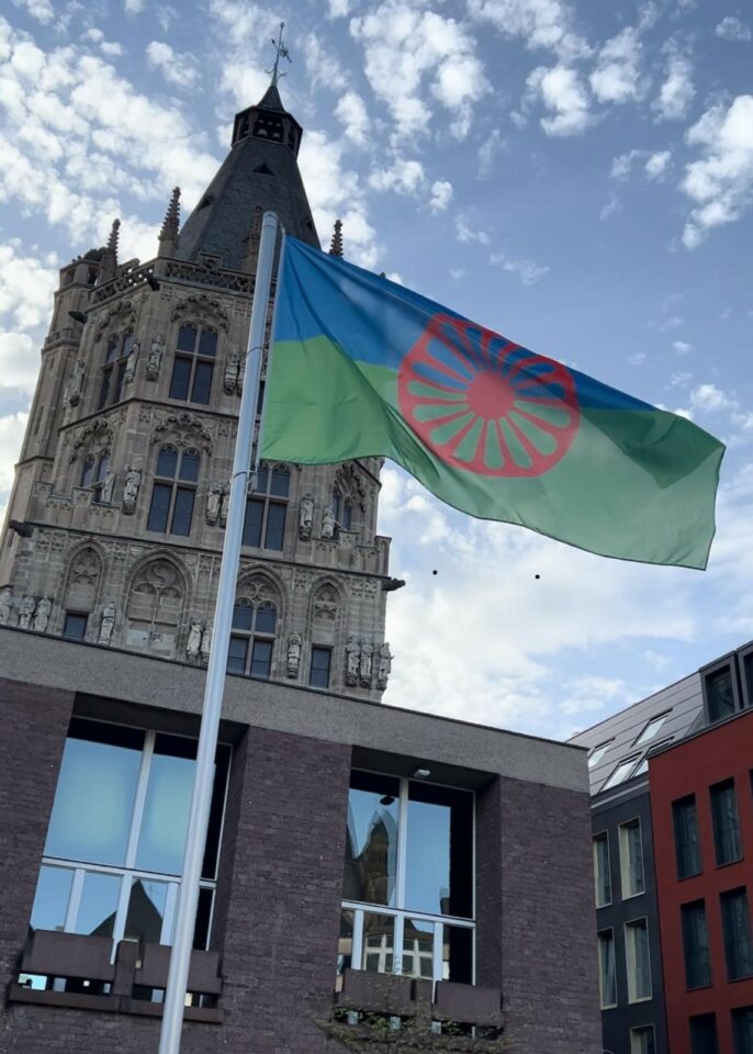 Eine romanische Flagge mit blauen und grünen Feldern und einem roten Rad weht an einem Fahnenmast vor einem historischen europäischen Gebäude, mit einem bewölkten Himmel im Hintergrund.