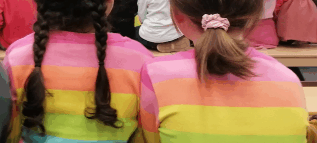 Zwei Kinder sitzen nebeneinander auf Bänken, von hinten gesehen. Beide tragen Pullover mit horizontalen Regenbogenstreifen. Ein Kind hat geflochtene Haare, das andere einen Pferdeschwanz mit einem rosa Haargummi. Vor den beiden sitzen Menschen.